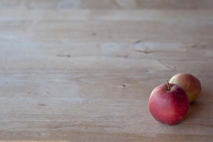Two red apples are clustered in one corner of a wooden table, drawing attention to the fruit in an open space. 