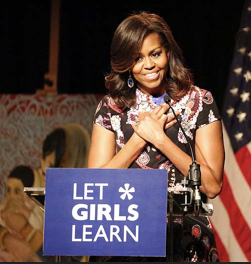 An image of Michelle Obama smiling as she delivers a speech from behind a sign that says "let girls learn"