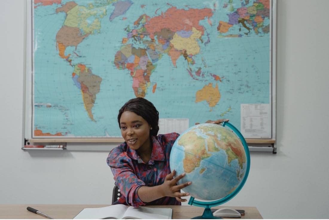Image of a woman witting at a desk and holding globe with world map on the wall behind her. 