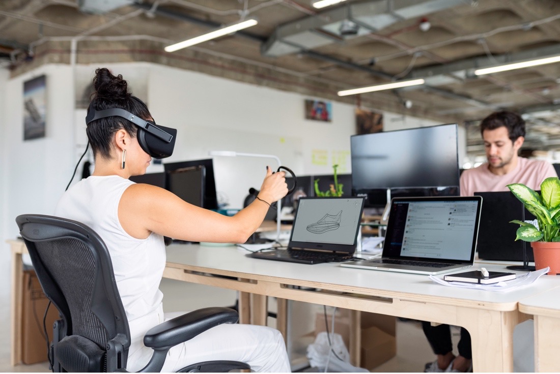 Image of a man and a woman sitting on opposite sides of a table, using multiple laptops. The woman is wearing a virtual reality headset. 